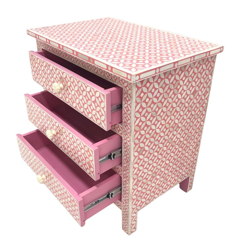 May include: A pink three-drawer nightstand with a geometric pattern. The drawers are open, revealing a pink interior. The knobs are cream-colored. The nightstand is made of wood and has a decorative design.