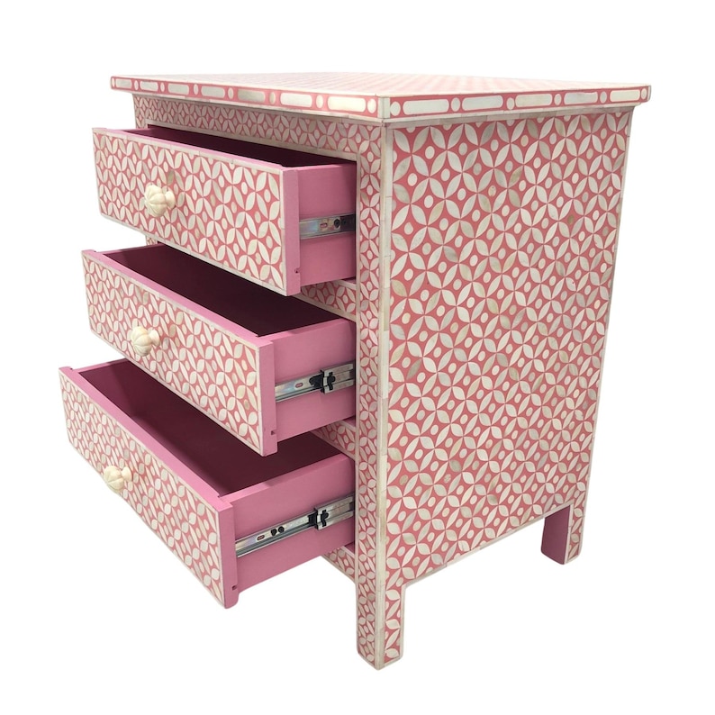 May include: A pink three-drawer nightstand with a geometric pattern. The drawers are open, revealing a pink interior. The knobs are off-white. The nightstand is approximately 24 inches tall.