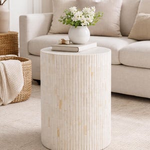 May include: A cylindrical side table with a textured, light-colored surface. The table is topped with a white vase holding white flowers, a book, and decorative objects. The table is in a living room setting with a sofa and rug.
