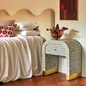 Könnte beinhalten: Schlafzimmer-Interieur mit Bett, weißer Bettwäsche und dekorativen Kissen. Ein gestreifter Nachttisch mit Schublade und goldenen Akzenten steht neben dem Bett. Eine Blumenvase und ein abstraktes Kunstwerk ergänzen die Einrichtung.