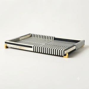 May include: A rectangular tray featuring a high-contrast black and white striped pattern arranged in alternating horizontal and vertical sections. The tray is supported by four small, metallic gold-toned feet at each corner of the base.