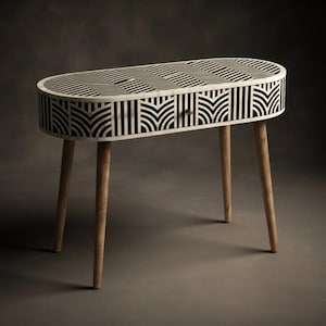 May include: An oval console table featuring a black and white geometric pattern across the top and front drawer. The piece is supported by four light-toned wooden legs and includes a single small, round, metallic-colored drawer knob.