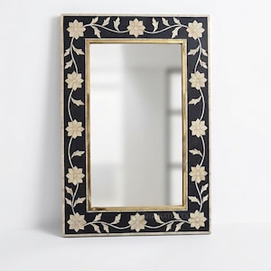 Puede incluir: Un espejo de pared rectangular con un marco negro y un patrón de enredadera floral blanca incrustado. El espejo tiene un borde interior fino de tono dorado que rodea la superficie de cristal reflectante, creando un aspecto limpio.