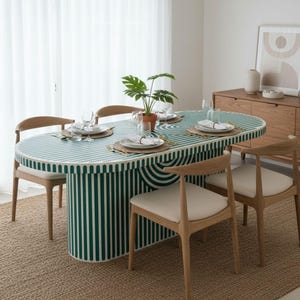 Puede incluir: Una mesa de comedor ovalada con una parte superior y una base a rayas verdes y blancas. La mesa está rodeada por cuatro sillas de madera con asientos de color crema. Una planta en maceta se encuentra en el centro de la mesa, que está puesta con platos, vasos y cubiertos.