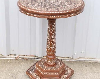 May include: A handcrafted, brown wooden side table with intricate inlaid patterns. The round tabletop, pedestal, and hexagonal base feature detailed designs in a lighter color. The table stands approximately 28 inches tall.