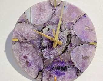 May include: Circular wall clock with a natural amethyst stone face. The clock has gold-coloured hands and hour markers. The amethyst features shades of purple, white, and black. A decorative item for home decor.