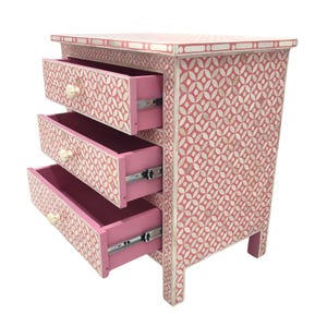 May include: A pink three-drawer chest with a geometric pattern in white. The drawers are open, revealing a pink interior. The knobs are off-white. The chest is approximately 30 inches tall.