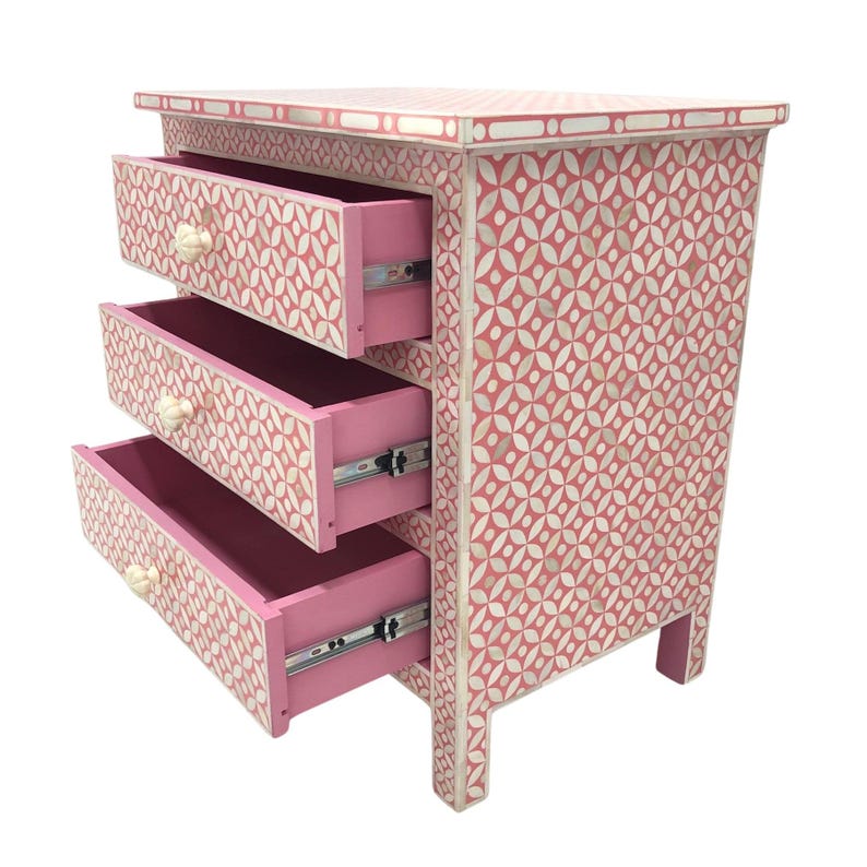 May include: A pink three-drawer chest with a geometric pattern in white. The drawers are open, revealing a pink interior. The knobs are off-white. The chest is approximately 30 inches tall.