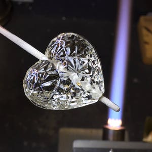 Puede incluir: Un corazón de vidrio transparente con un patrón detallado se está calentando con un soplete. El corazón está suspendido en una varilla de metal.