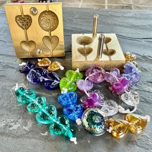 Puede incluir: Dos moldes de latón para hacer cuentas de vidrio. Los moldes tienen forma de corazones y flores. Una variedad de cuentas de vidrio de colores hechas con los moldes están dispersas alrededor de los moldes.