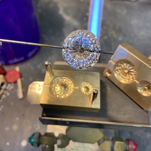 Könnte beinhalten: Eine silberne Metallperle mit einem detaillierten Blumenmuster wird mit einer Fackel erhitzt und von einer Metallstange über einer Messingform gehalten.