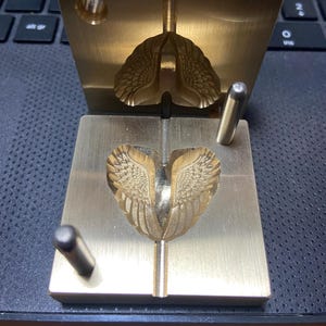 Può includere: Uno stampo in ottone per l'artigianato, con un design dettagliato ad ali. Lo stampo è aperto, rivelando l'intricato motivo ad ali scolpito nel metallo. Sono visibili due perni cilindrici, insieme a un manico in legno.