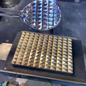 Puede incluir: Un objeto de vidrio transparente con un patrón geométrico texturizado es sostenido por una herramienta de metal. El objeto se coloca sobre una placa de metal cuadrada dorada con un diseño de pirámide repetitivo. Una llama azul es visible en el fondo.