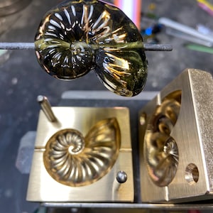 May include: A glass nautilus shell being heated over a torch, with a brass mould in the background. The shell is a deep green colour with gold accents.