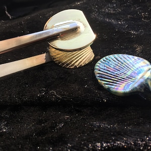 May include: Two decorative metal seashells, one gold and one iridescent blue, on a black fabric background.