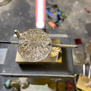 May include: A clear glass object with a textured, radial pattern is being heated with a torch. The object is held in place by a metal stand.