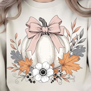 Op de afbeelding: Crèmekleurige sweatshirt met een wit pompoenontwerp, versierd met een roze strik en herfstbladeren. Het ontwerp bevat oranje en grijze bladeren, een witte bloem en bessen, wat een seizoensgebonden herfstthema creëert.