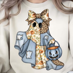 May include: A cream-colored sweatshirt featuring a whimsical ghost design. The ghost is adorned with a patchwork design of floral and denim patterns, a leopard print bow, and a jack-o'-lantern bucket. The design is perfect for Halloween.