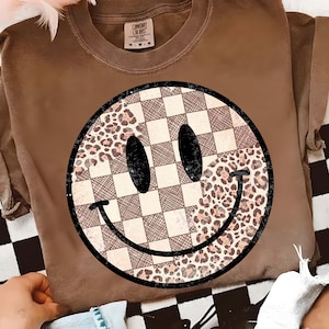 May include: Brown t-shirt with a large smiley face design. The smiley face combines checkered, leopard print, and solid black elements. The shirt has rolled-up sleeves and a crew neck.