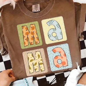 May include: Brown t-shirt with the word "MAMA" in a patchwork design. Each letter is in a different square with various patterns, including pumpkins, leaves, and floral designs. The shirt has a vintage, worn look.