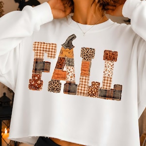 May include: A white long-sleeve sweatshirt with a raw-edge hem featuring the word FALL in large block letters. The letters are composed of various fabric patches in brown, orange, and tan tones, including plaid, leopard print, and floral patterns.