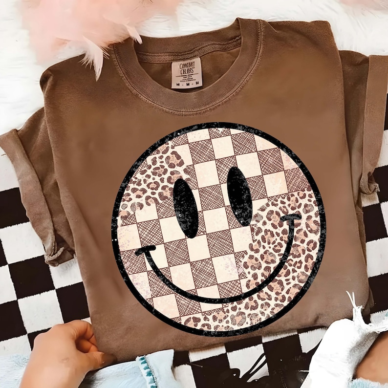 以下が含まれることがあります： 大きなスマイリーフェイスのデザインが特徴の茶色のTシャツ。スマイリーフェイスは、チェック柄、ヒョウ柄、無地の黒の要素を組み合わせています。シャツは袖をまくり、クルーネックです。