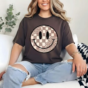 以下が含まれることがあります： スマイリーフェイスのデザインが施された濃い茶色のTシャツ。スマイリーフェイスは、片側にチェック柄、もう片側にヒョウ柄が施されています。目は楕円形で、口は曲線です。