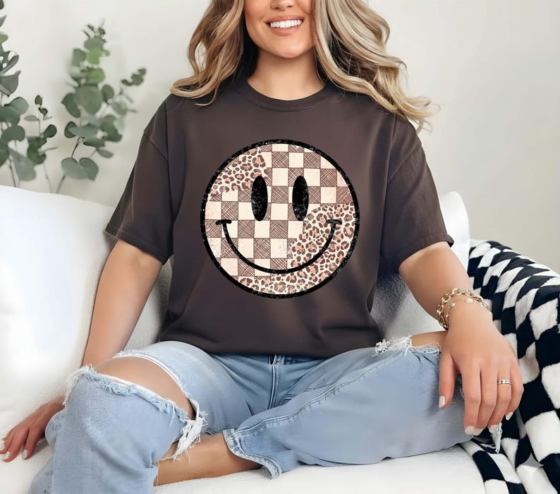 以下が含まれることがあります： スマイリーフェイスのデザインが施された濃い茶色のTシャツ。スマイリーフェイスは、片側にチェック柄、もう片側にヒョウ柄が施されています。目は楕円形で、口は曲線です。