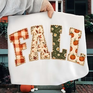 Peut inclure: Un t-shirt blanc avec le mot FALL écrit en grandes lettres majuscules. Le F présente un motif à carreaux rouge et blanc, le A un imprimé de feuilles d'automne, le L est vert foncé avec des marguerites blanches et le L a un motif de citrouilles.