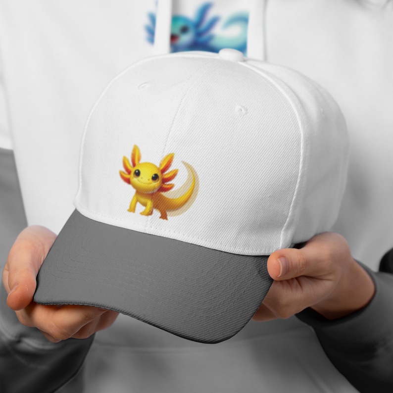 Puede incluir: Gorra de b&eacute;isbol blanca con visera gris, adornada con un ajolote de dibujos animados en amarillo y naranja. El ajolote tiene una cara sonriente y branquias detalladas. La gorra la sostiene una persona.