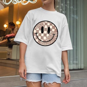 以下が含まれることがあります： 大きなスマイリーフェイスのデザインが特徴の白いTシャツ。スマイリーフェイスは、市松模様とヒョウ柄のセクションに分かれています。このシャツは半袖で、ゆったりとしたフィット感です。