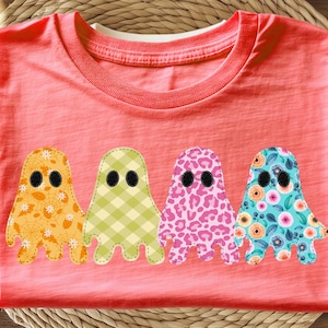 May include: Coral-colored t-shirt featuring four whimsical ghost appliqués. Each ghost has a unique pattern: orange floral, green gingham, pink leopard print, and blue floral. The ghosts have black oval eyes and are stitched onto the shirt.