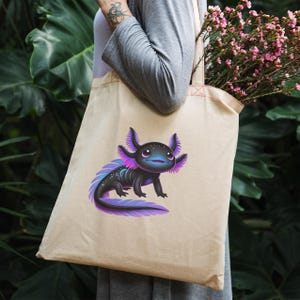 Puede incluir: Bolso tote beige con una ilustraci&oacute;n de dibujos animados de un ajolote. El ajolote es negro con detalles morados y rosas. El bolso tote lo lleva una persona con una camisa gris de manga larga. El fondo es verde exuberante.