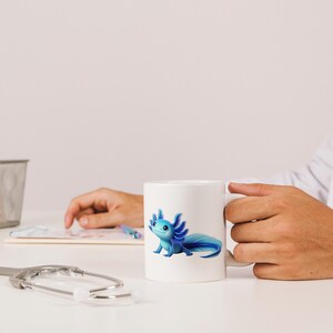 Puede incluir: Taza de cer&aacute;mica blanca con una ilustraci&oacute;n de ajolote azul. El ajolote tiene branquias detalladas y una cola larga. La taza est&aacute; sostenida por una persona. El dise&ntilde;o es vibrante y llamativo.