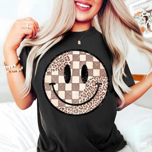 以下が含まれることがあります： 大きなスマイリーフェイスのデザインが特徴の黒いTシャツ。スマイリーフェイスは2つのセクションに分かれており、片方は市松模様、もう片方はヒョウ柄です。デザインは黒で縁取られています。