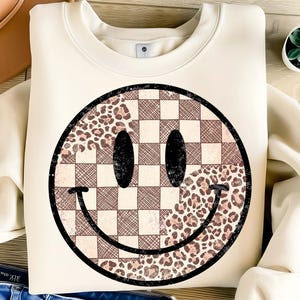 以下が含まれることがあります： クリーム色のスウェットシャツ。大きなスマイリーフェイスのデザイン。スマイリーフェイスは、茶色とタンの色合いのチェック柄、ヒョウ柄、クロスハッチパターンを組み合わせています。目と口は黒です。