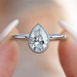 May include: A pear-shaped diamond ring with a halo setting. The ring features a large, clear diamond with a brilliant cut, set in a delicate silver band. The halo is made of small, sparkling stones. The ring is held between two fingers.