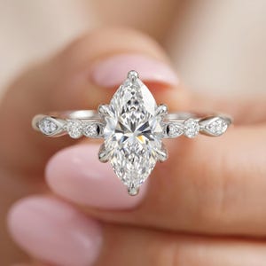 Op de afbeelding: Een zilverkleurige ring met een grote marquise-geslepen middensteen die door klauwen wordt vastgehouden. De band is aan weerszijden versierd met kleinere ronde en marquisevormige stenen, wat zorgt voor een symmetrisch ontwerp met een gepolijste afwerking.