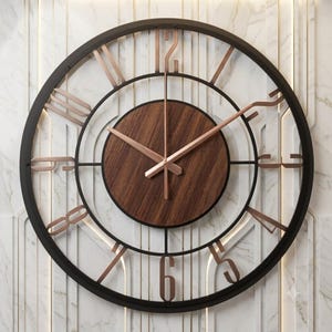 May include: A large circular wall clock featuring a dark metal outer frame and a central circular wooden dial with a visible grain. The clock has copper-coloured metal numerals and matching slender hands, creating a contrast against the dark frame.