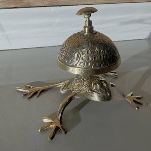 Könnte beinhalten: Eine glockenförmige Messingglocke in Form eines Frosches mit einem kuppelförmigen Oberteil und einem kleinen Griff. Die Glocke hat komplizierte Blumenmuster und besteht aus einem glänzenden, metallischen Material. Die Beine des Frosches sind ausgestreckt.