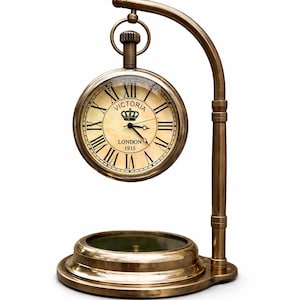 May include: A brass pocket watch clock with Roman numerals, the text "VICTORIA LONDON 1915" is visible. The clock is suspended from a curved stand with a circular base, creating a classic, antique aesthetic.