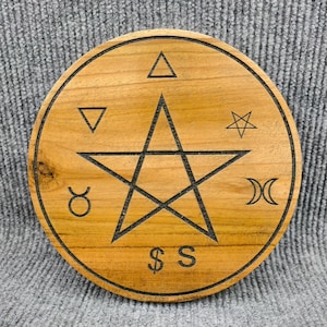 May include: A round wooden coaster with a pentagram and various symbols etched into the surface. The coaster is made of light brown wood and features a black outline and symbols. The symbols include a triangle, inverted triangle, star, crescent moon, and dollar sign.