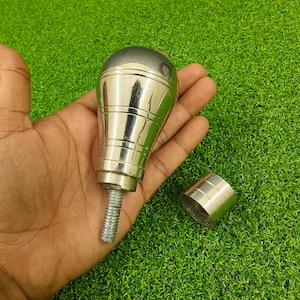 以下が含まれることがあります： 丸みを帯びた上部とねじ付きベースを備えた、光沢のあるシルバー色のギアシフトノブ。 別途、お揃いのシルバーリングが隣にあります。 ノブは、緑色の背景に対して手で保持されています。