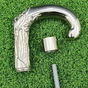 May include: A silver walking cane handle with a tree design on the grip. The curved handle, a separate metal piece, and a screw are visible. The cane handle is on a green surface.