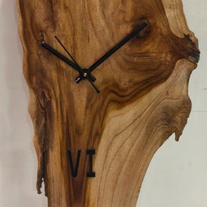 Könnte beinhalten: Eine Wanduhr aus Holz mit natürlicher Kante. Das Zifferblatt besteht aus einem einzigen Holzstück mit sichtbarer Maserung und Textur. Schwarze römische Ziffern markieren XII und VI. Schwarze Zeiger.