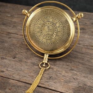 Puede incluir: Un colgante circular de color dorado con intrincados grabados, incluyendo números romanos y símbolos. El colgante está suspendido de una cadena dorada a juego con una borla. El diseño sugiere un estilo vintage o antiguo, posiblemente una brújula o una herramienta de navegación.