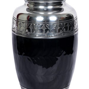 May include: A decorative urn with a black body and silver accents. The urn features a silver lid and base with floral patterns. The body has a textured, glossy finish. The urn is designed for memorial purposes.