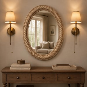 May include: An oval wall mirror with a thick, braided rope frame. The mirror is flanked by two brass wall sconces with beige fabric shades. Below the mirror sits a wooden console table with three drawers, two stacked books, and a rectangular tray.