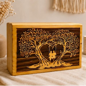Può includere: Scatola di legno con un disegno intagliato di due alberi che formano un cuore, con una coppia su un'altalena. La scatola ha una cornice in legno chiaro e un pannello in legno scuro con il disegno intagliato. La scatola è lunga circa 20 cm.