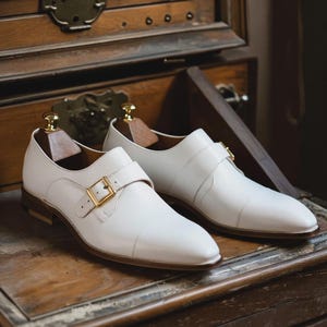 May include: A pair of white leather dress shoes with a buckle closure. The shoes have a gold-colored buckle and a brown sole. The shoes are displayed on a wooden surface.
